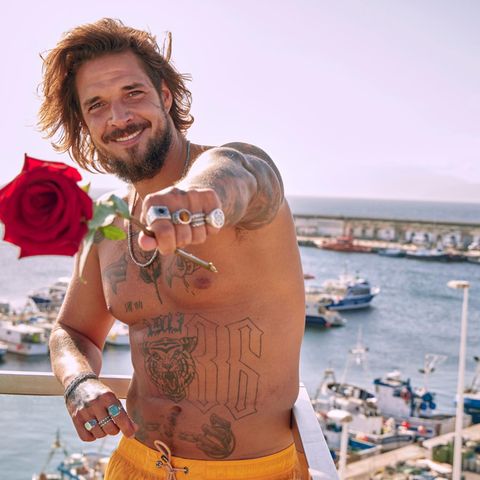 Zico Banach posiert oberkörperfrei an einem Strand. Er grinst breit und hält mit der linken Hand eine Rose in die Kamera.